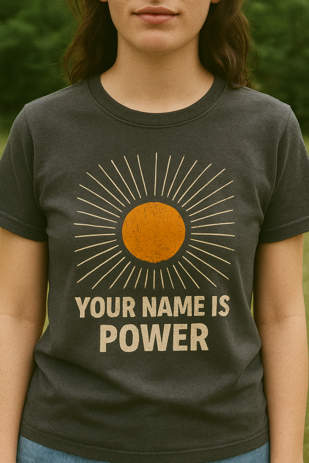 woman standing in a park wearing a T-shirt with a distressed orange sun graphic radiating white rays, above bold text that reads: "YOUR NAME IS POWER." Front, Charcoal