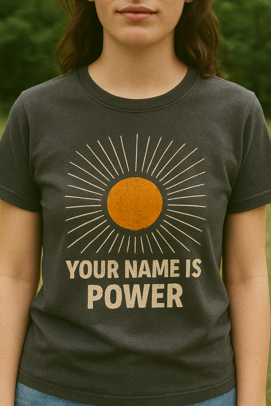 woman standing in a park wearing a T-shirt with a distressed orange sun graphic radiating white rays, above bold text that reads: "YOUR NAME IS POWER." Front, Charcoal