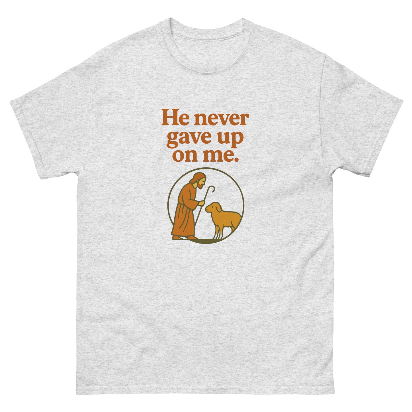 Christian T-shirt with the phrase 'He never gave up on me' above an illustration of Jesus as the shepherd standing with a sheep inside a circular frame. Front, Ash color