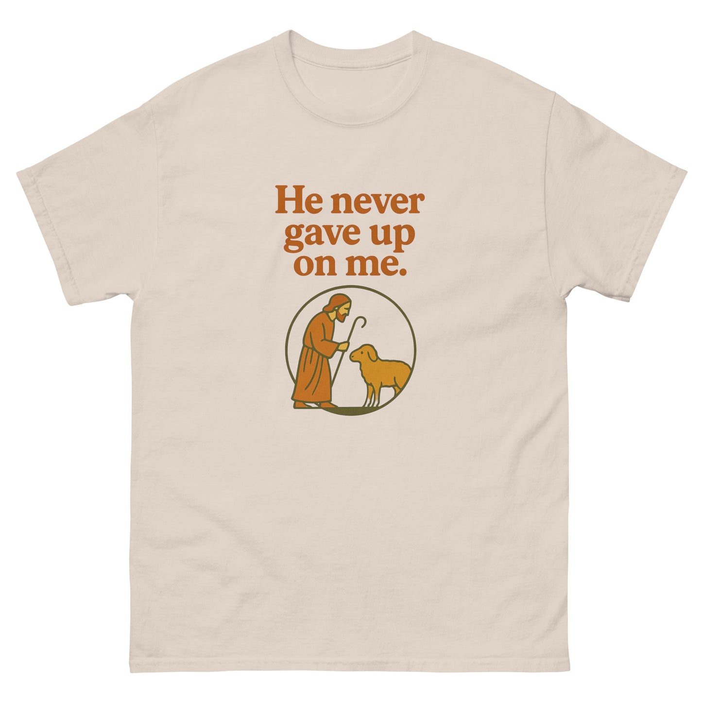 Christian T-shirt with the phrase 'He never gave up on me' above an illustration of Jesus as the shepherd standing with a sheep inside a circular frame. Front, natural color