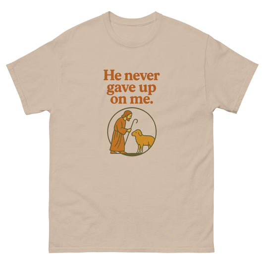 Christian T-shirt with the phrase 'He never gave up on me' above an illustration of Jesus as the shepherd standing with a sheep inside a circular frame. front, sand color