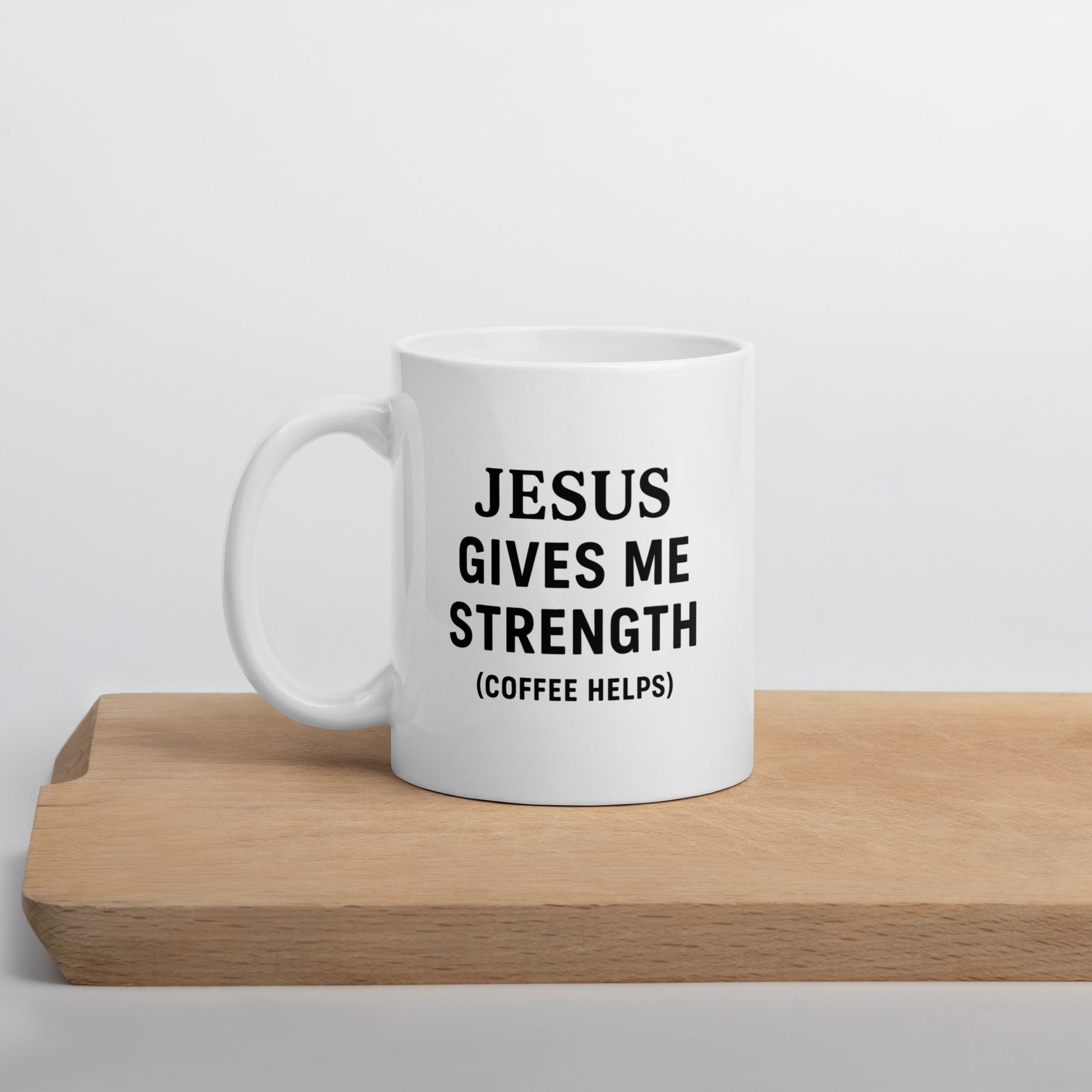 white coffee mug that says 'JESUS GIVES ME STRENGTH (COFFEE HELPS)' sitting on a cutting board in a light white background