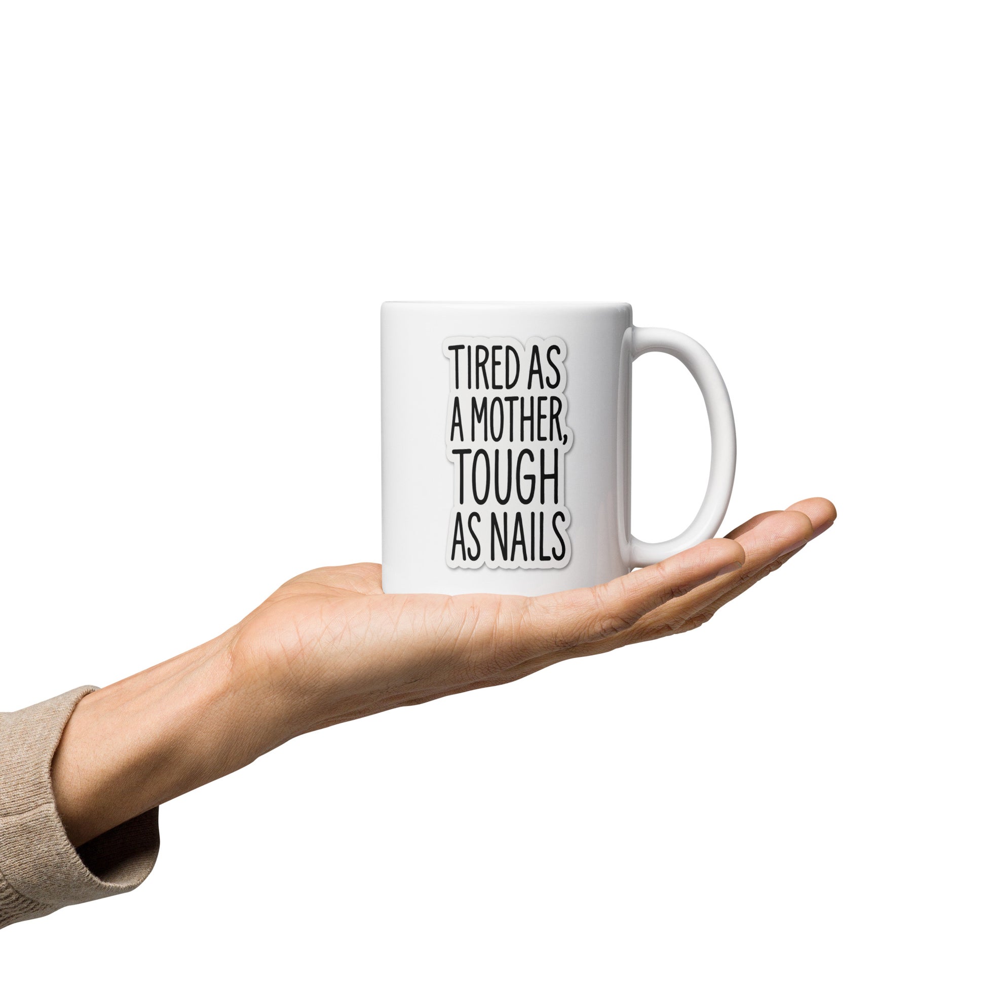 Hand holding a white coffee mug with bold black text that says 'Tired As A Mother, Tough As Nails' against a clean white background.